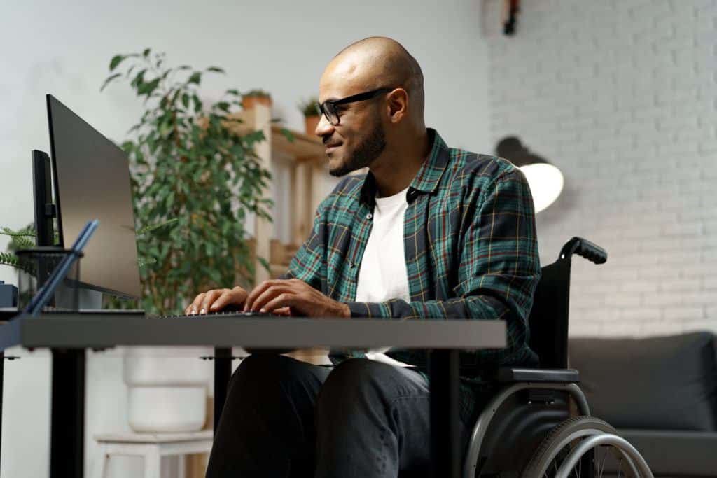 Disabled young african american man in wheelchair using computer while sitting at his working table. people with disabilities
