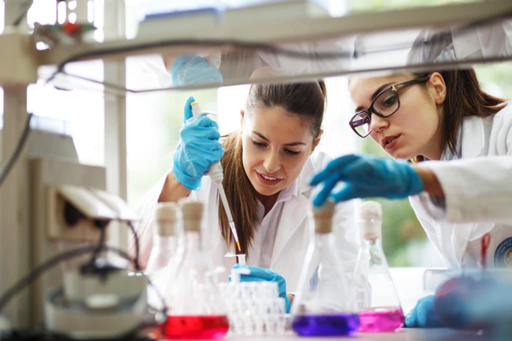 two women in lab coats and gloves looking at something
